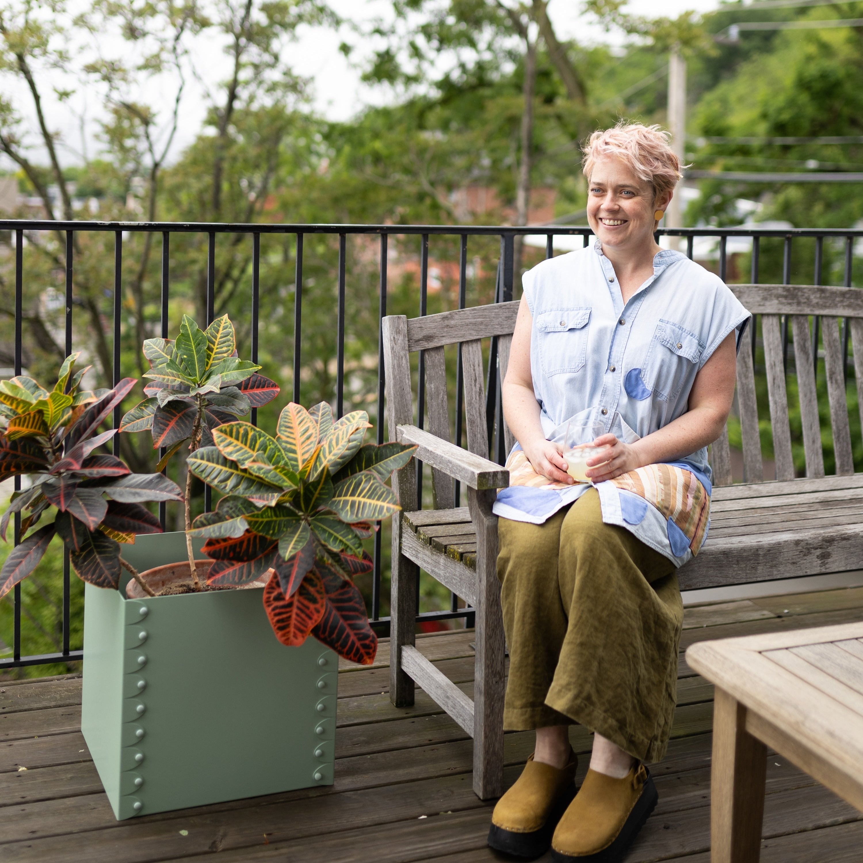 Person sitting on a wooden bench with plants and a table in an outdoor setting